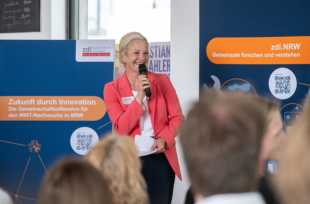 Eine Frau in einem rosa Blazer spricht bei einer Veranstaltung in ein Mikrofon, hinter ihr hängen blaue Banner mit Aufschriften zu den Themen Innovation und MINT-Bildung.