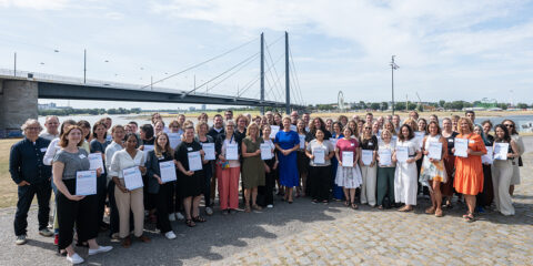 Eine große Gruppe steht im Freien an einem Fluss und hält bei der zdi-Qualitätssiegelübergabe 2026 Urkunden in den Händen. Im Hintergrund sind eine Brücke und eine Stadtlandschaft zu sehen.