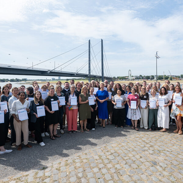 Eine große Gruppe steht im Freien an einem Fluss und hält bei der zdi-Qualitätssiegelübergabe 2026 Urkunden in den Händen. Im Hintergrund sind eine Brücke und eine Stadtlandschaft zu sehen.