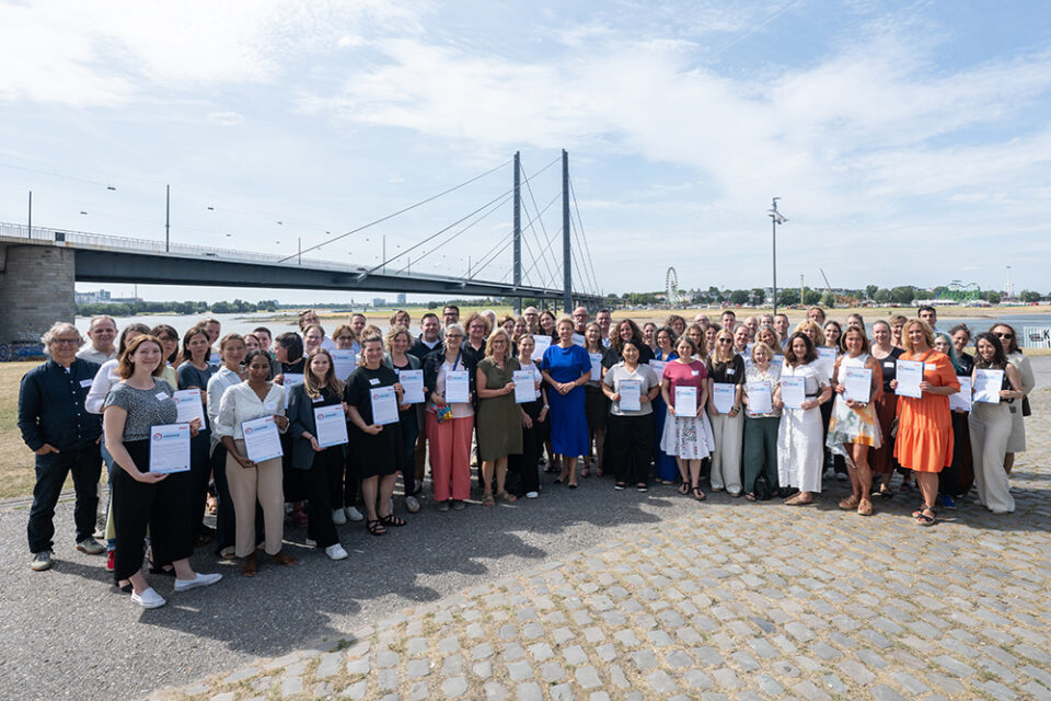 Eine große Gruppe steht im Freien an einem Fluss und hält bei der zdi-Qualitätssiegelübergabe 2026 Urkunden in den Händen. Im Hintergrund sind eine Brücke und eine Stadtlandschaft zu sehen.