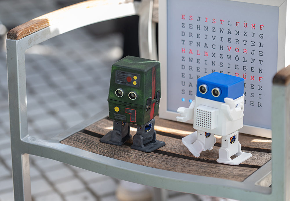 Ein grüner und ein weißer Spielzeugroboter stehen auf einem Holzstuhl neben einem gerahmten Wortsuchrätsel mit roten und schwarzen Buchstaben.