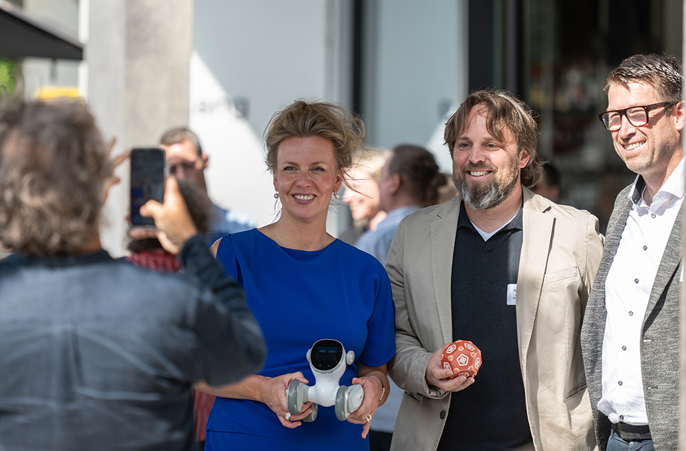 Drei Personen posieren im Freien bei der Verleihung des zdi-Qualitätssiegels 2026. Eine hält einen Roboter in der Hand, eine andere zeigt auf einen roten Gegenstand.