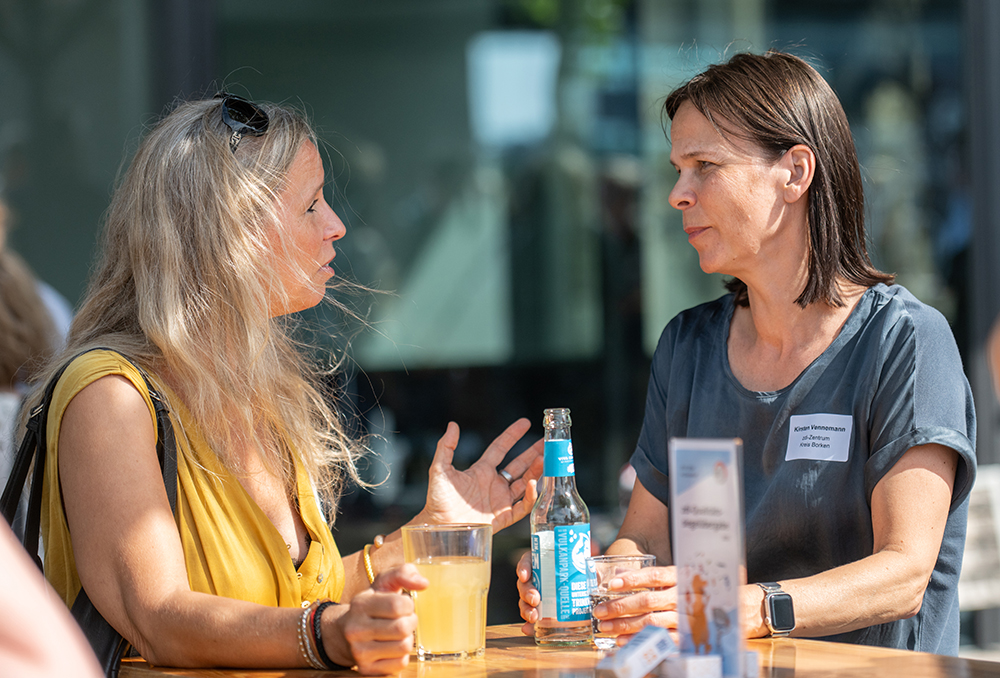 Zwei Frauen sitzen an einem Tisch im Freien und unterhalten sich. Die eine trinkt Saft, die andere Sprudelwasser. Ein Namensschild an einer der beiden weist auf die zdi-Veranstaltung 2026 hin.