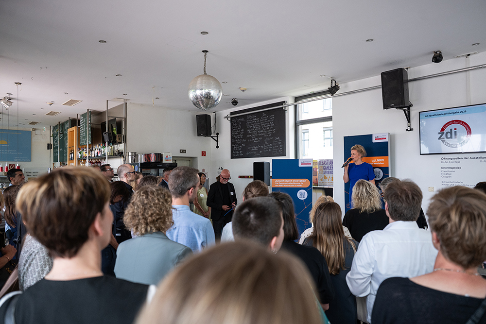 Eine Frau spricht von einem Podium aus zu den Zuhörern in einem hell erleuchteten Raum mit einer Discokugel, einem Barbereich und Informationsbannern.