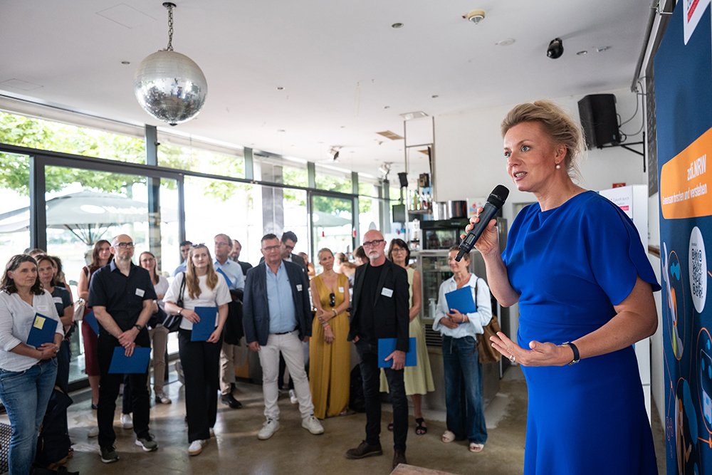 Eine Frau in einem blauen Kleid spricht bei der zdi-Qualitätssiegelübergabe 2026 in der Halle vor einem aufmerksamen Publikum ins Mikrofon.