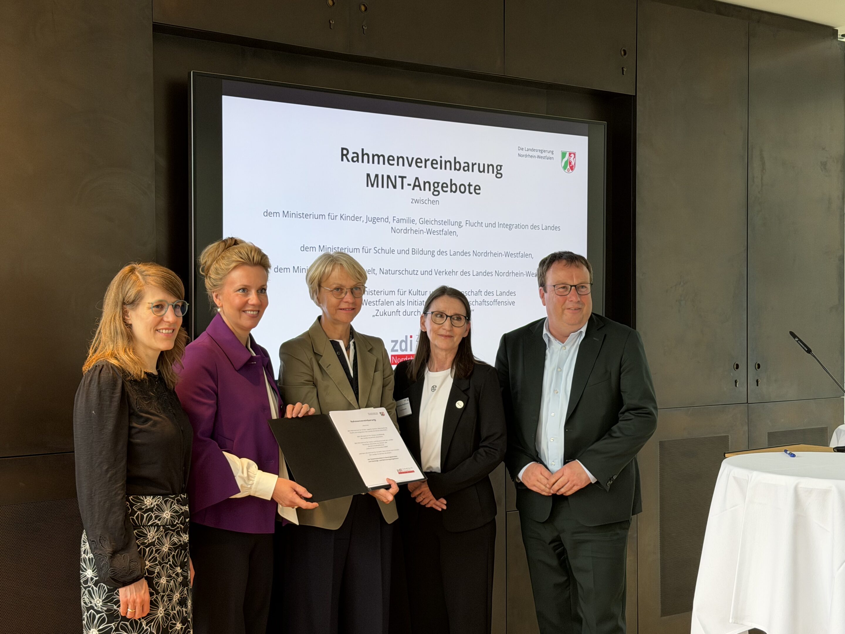 Fünf Personen in Geschäftskleidung stehen zusammen und halten eine Urkunde in den Händen. Hinter ihnen ist auf einer Leinwand eine Präsentationsfolie mit dem Titel „Rahmenvereinbarung MINT-Angebote“ zu sehen, die die Bedeutung von MINT in der Ganztagsgrundschule hervorhebt.
