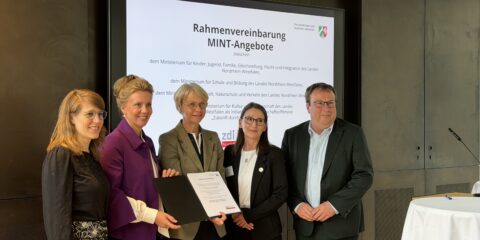 Fünf Personen in Geschäftskleidung stehen zusammen und halten eine Urkunde in den Händen. Hinter ihnen ist auf einer Leinwand eine Präsentationsfolie mit dem Titel „Rahmenvereinbarung MINT-Angebote“ zu sehen, die die Bedeutung von MINT in der Ganztagsgrundschule hervorhebt.