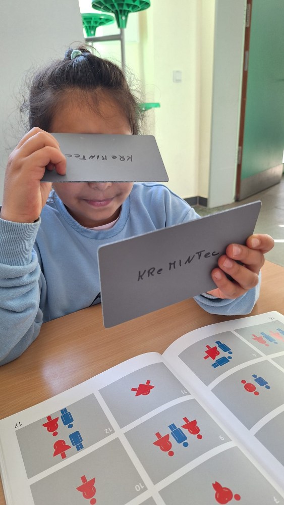 Ein Kind sitzt an einem Schreibtisch und hält zwei rechteckige Karten mit Schriftzug hoch. Das Kind schaut in ein aufgeschlagenes Buch, in dem bunte Symbole und Zahlen zu sehen sind.