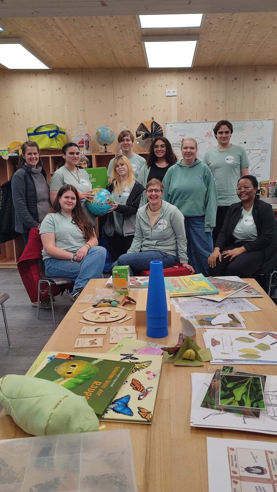 Zehn Erwachsene posieren in einem Klassenzimmer für ein Foto. Auf dem Tisch vor ihnen liegen Naturwissenschaftsbücher, ein Globus und Unterrichtsmaterialien.